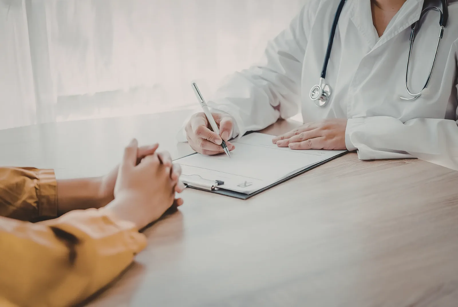 A consultation in progress: one person writes on a document while the other sits opposite with their hands clasped together on the table.
