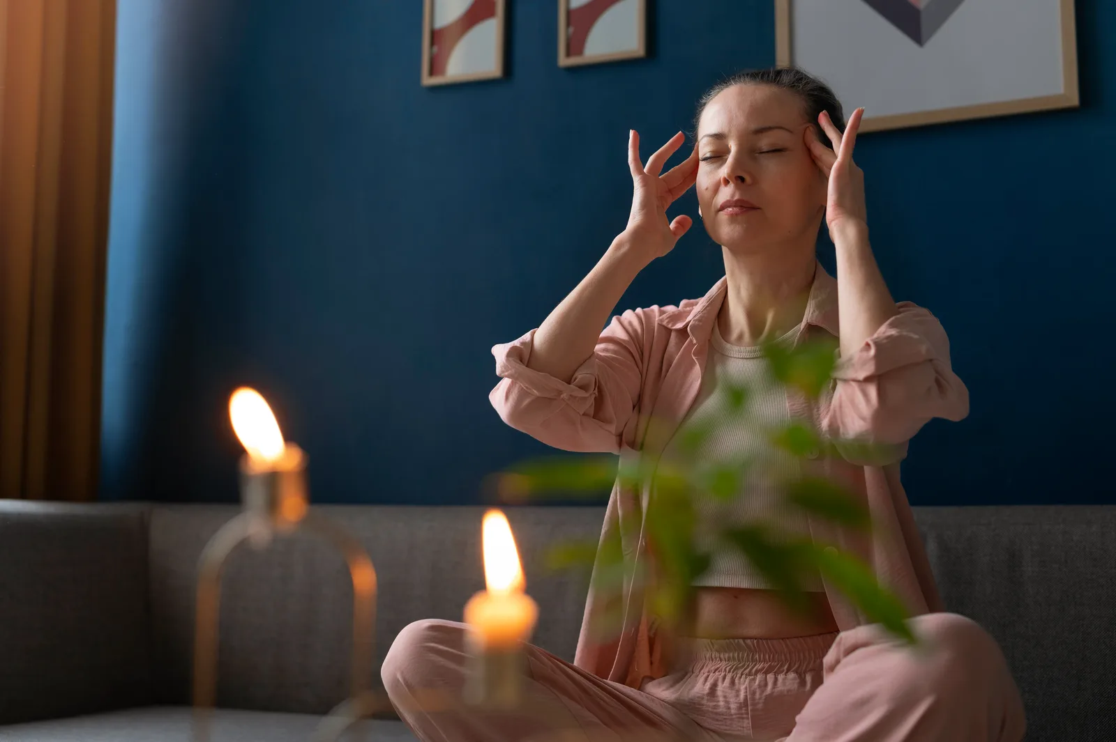 A woman sits cross-legged on a sofa with her eyes closed and her fingertips resting at her temples, in a calm room lit by candlelight.