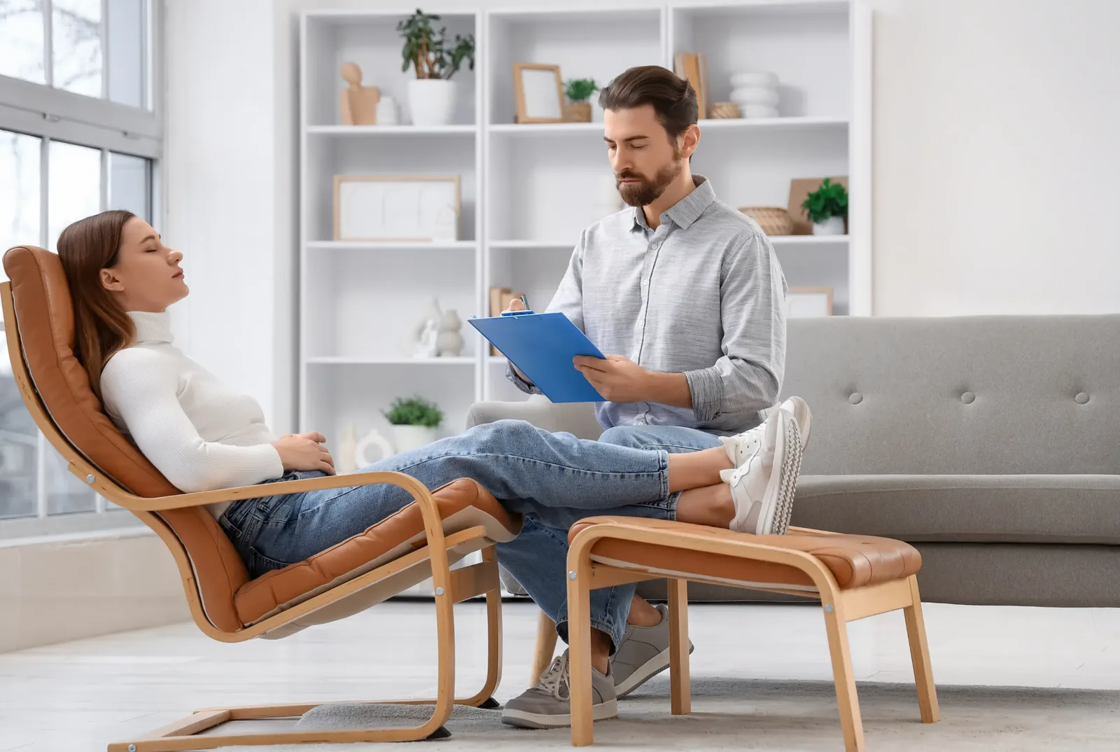 A practitioner holding a clipboard sits opposite a client who is resting back in a wooden reclining chair in a bright room.