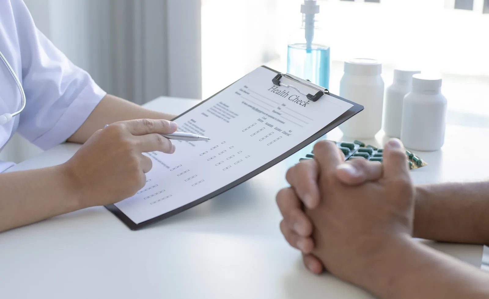 A healthcare professional in a white coat holds a health check form on a clipboard while seated across from a person with their hands clasped.