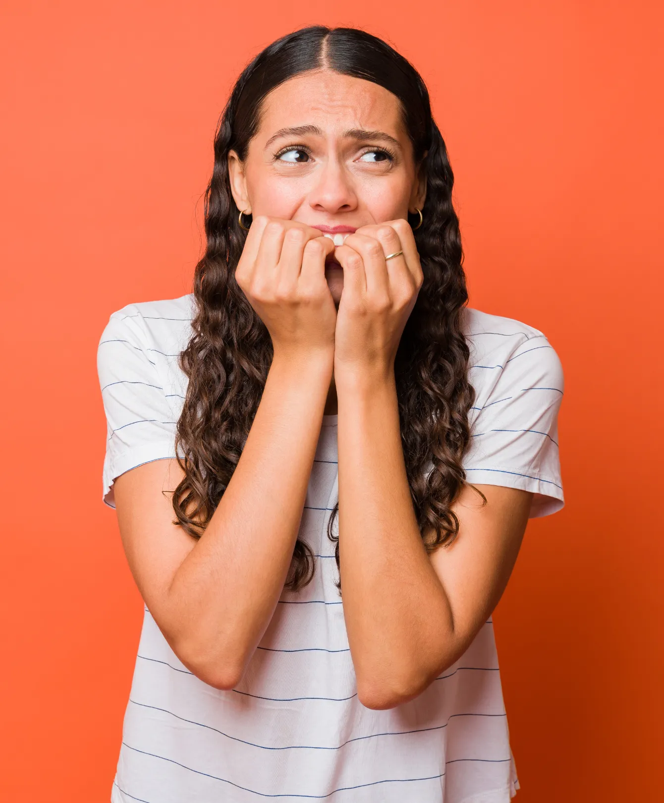 A woman with a tense, worried expression rests her closed hands against her mouth while looking away, photographed against a plain orange background.