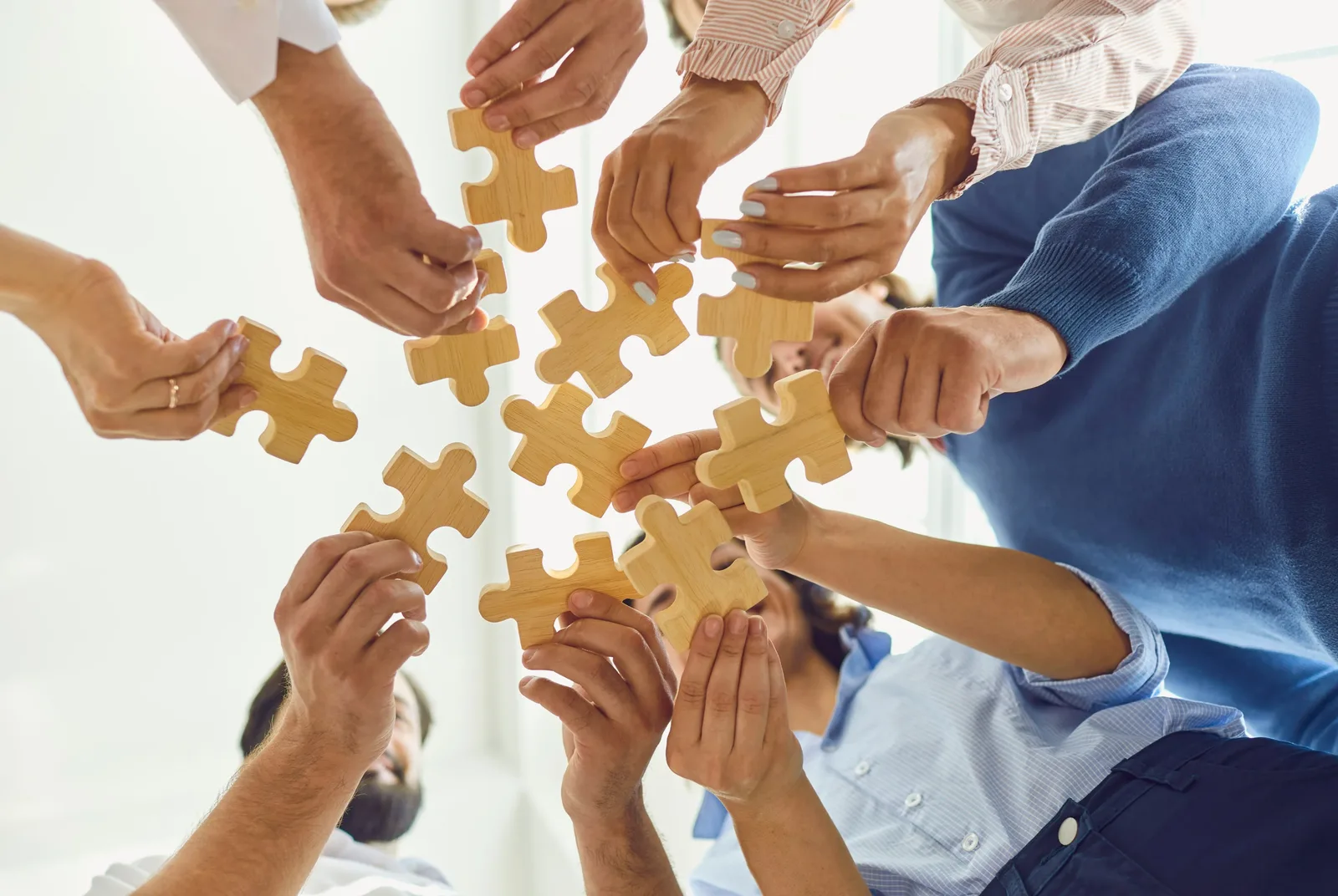 Several people stand in a circle, each holding a wooden jigsaw piece up towards the centre.